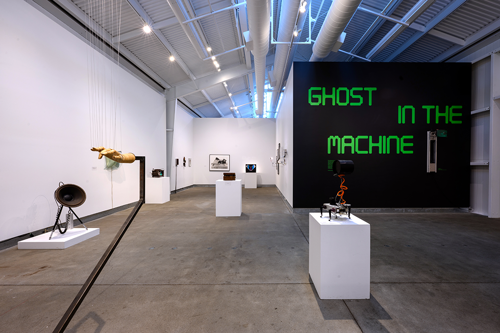 Various sculptures and wall works in a gallery, with wall text that reads "Ghost in the Machine" in green computer font