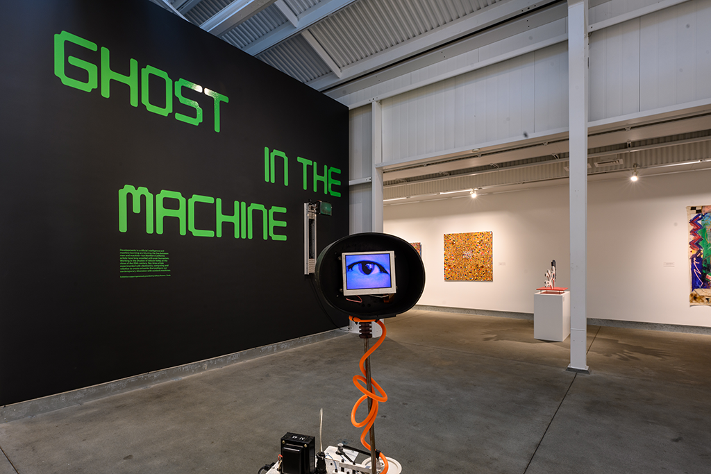 A sculpture with a video screen with an image of an eye, and a wall with "Ghost in the Machine" in large green computer type