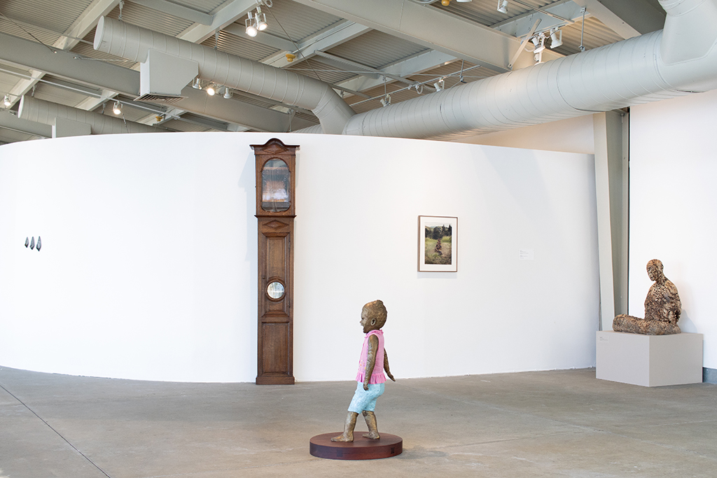 A gallery with a sculptural clock, a figurative sculpture of a child, and a photo on the wall