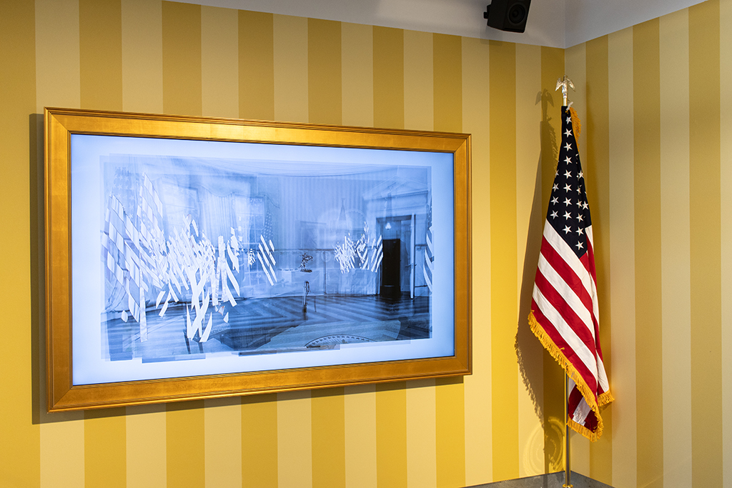 A video monitor with images from the oval office on a wall painted with yellow stripes