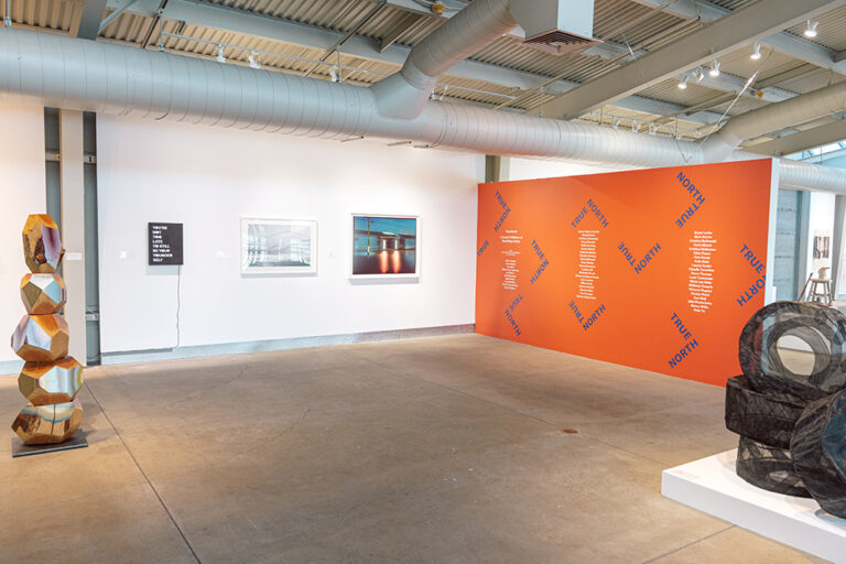 An assortment of artworks, including paintings, drawings, and sculpture, in a gallery with a large orange wall.