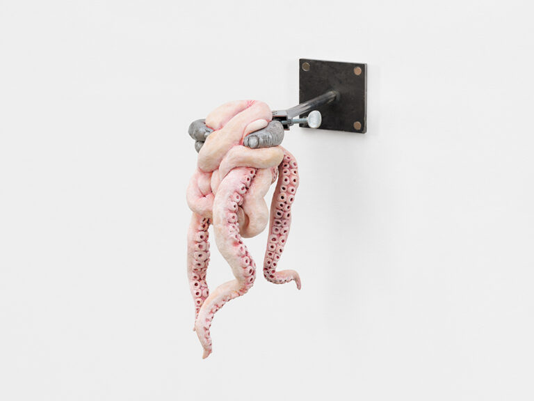 A sculpture of a mass of pink tentacles held by a metal vise grip in the form of human fingers, mounted to a wall.